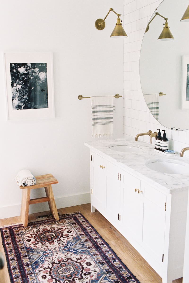 This all white modern vintage style bathroom is accented with brass and natural wood tones. A white double sink vanity has marble countertops and brass fixtures, A large circular mirror hangs on a white subway tile wall. The opposite wall is plain white with a single art print framed. A colorful ornate rug covers the wood floor.