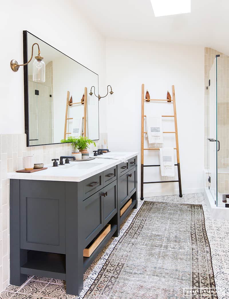 This modern vintage bathroom mixes a dark double sink vanity with white countertops with natural wood toles. Half the wall is tiled with sandy colored subway tiles. A large metal-framed rectangular mirror hangs above the large vanity. Next to the large glass walk-in shower is a ladder towel rack.