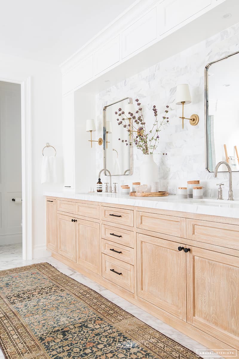 Another all-white modern vintage bathroom is balanced with a long natural wood vanity topped with marble countertops. The marble extends to the walls with a tiled backsplash inside a white cabinet alcove. Two silver framed mirrors hang above the double sinks, and a large oriental area rug covers the marble floor.