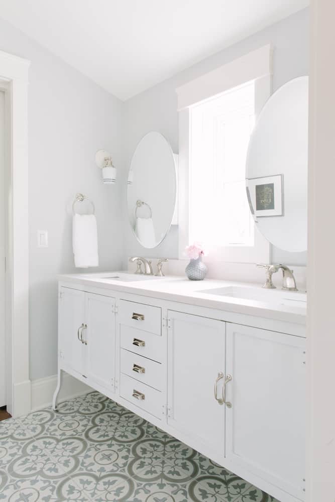 This bright and airy modern vintage style bathroom is all white with a double vanity with white cabinets and white marble countertop. The hardware is all brushed chrome, and the frameless oval mirrors are seamless with the rest of the design. The light gray walls echo the tones of the gray patterned tile floor.
