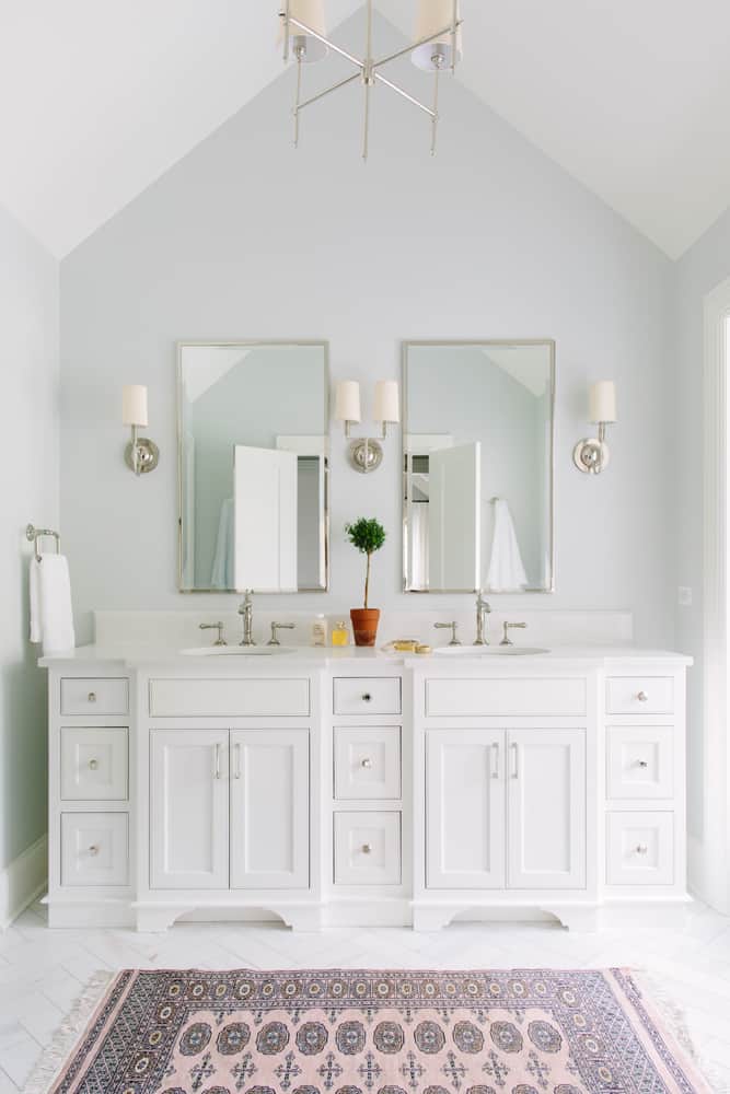 Another bright, all-white modern vintage style bathroom with a double vanity. The white cabinets and marble countertop of chrome hardware, and silver framed mirrors hang above the vanity. the marble tiled floor is covered with a light pink and blue oriental area rug.