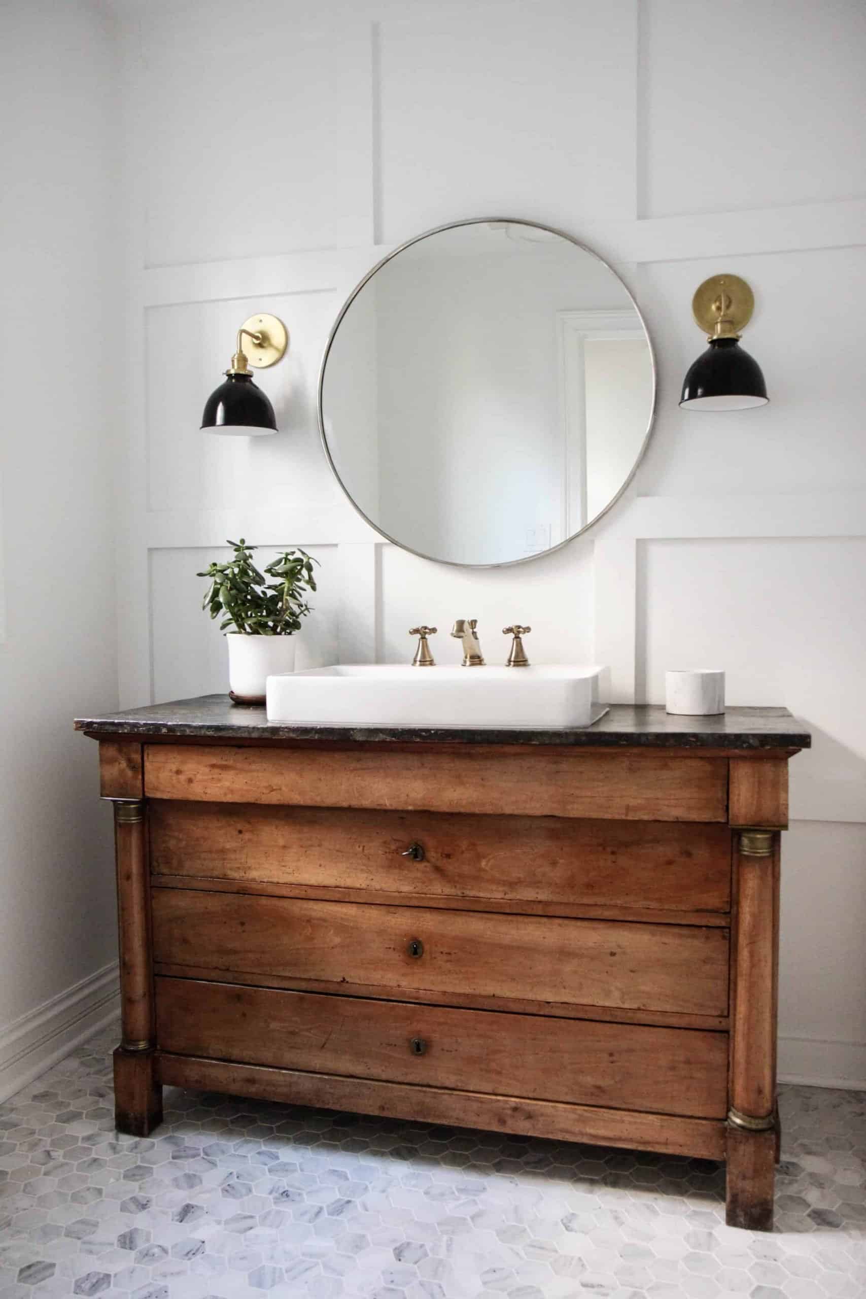 Another bathroom with hexagon floor times, these are marble tiles with white grout. The vintage vanity is a dark wood with drawers, and a white porcelain slink with brass faucets. A round mirror hangs between two vintage black and white wall sconces. The wall is covered in square wainscoting.