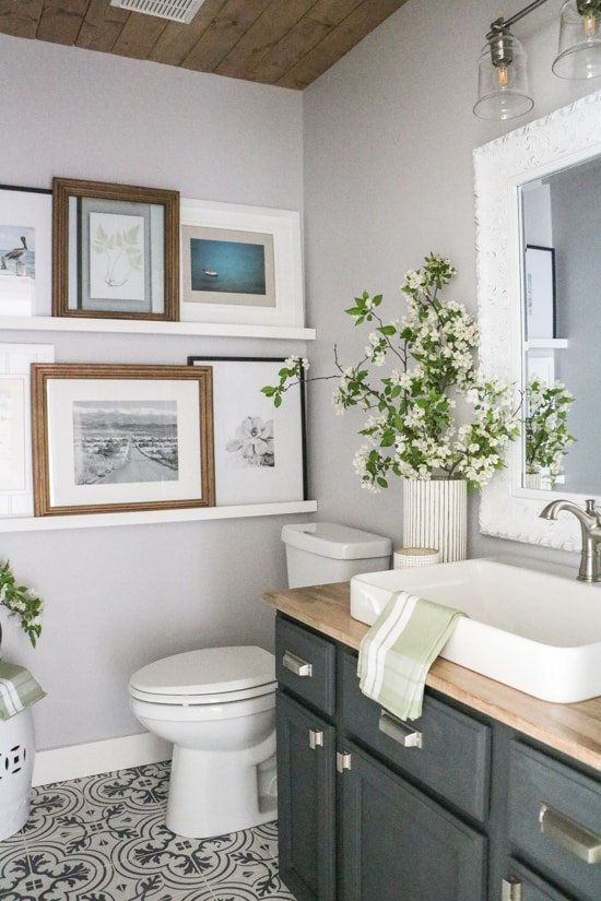 This modern vintage bathroom combines sleek style with personal touches. Against one wall are floating shelves, filled with framed artwork. A dark wood vanity had a white porcelain sink, with brushed chrome fixtures. A vintage white framed mirror hangs on the gray painted wall over the vanity.