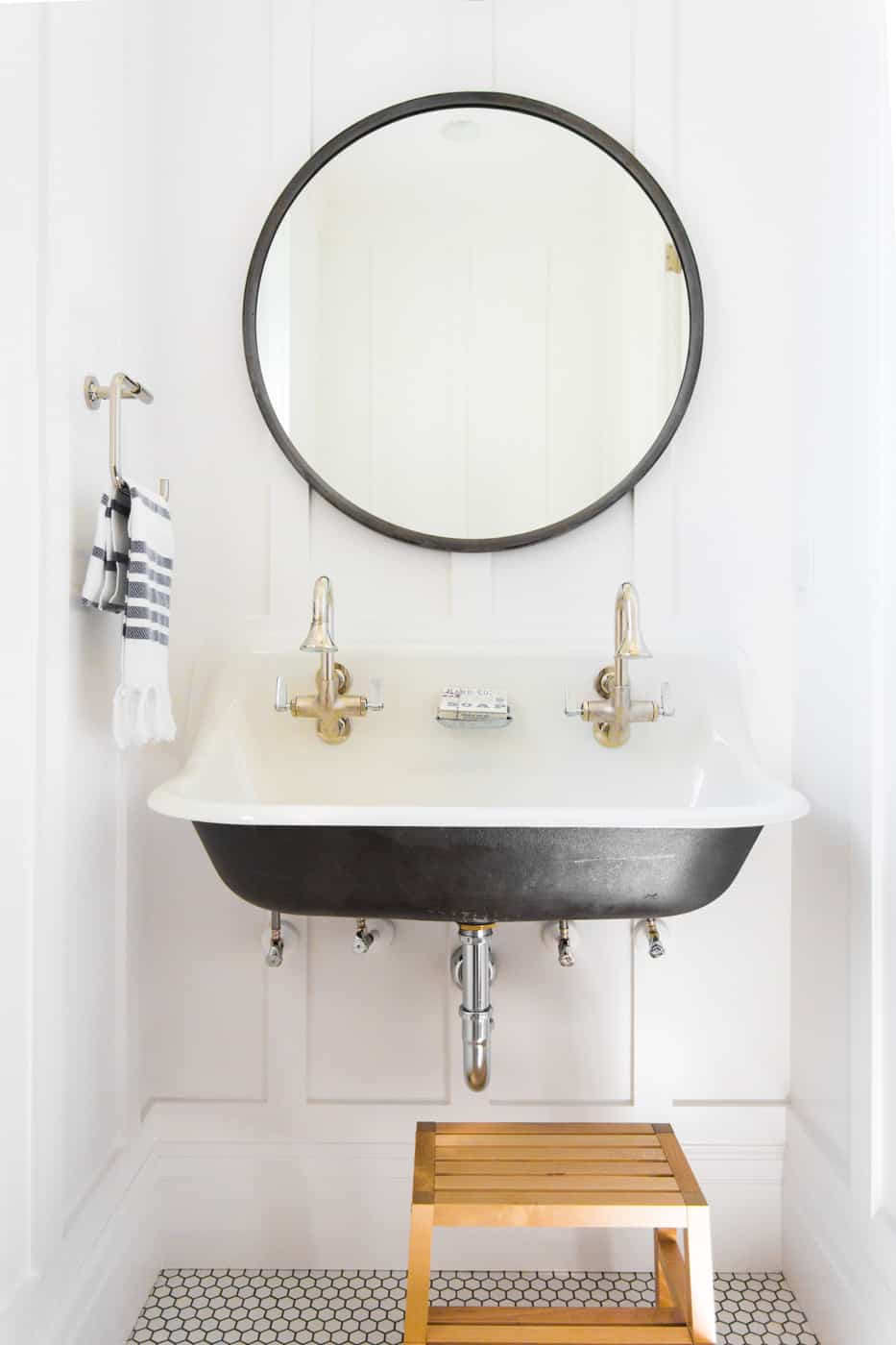 A deep sink is mounted to a white wainscoting covered wall, along with a round metal-framed mirror. The double faucets on the black and white sink are brass. A wooden step stool sits under the sink, and the floor is tiled with white hexagon tiles and dark grout.