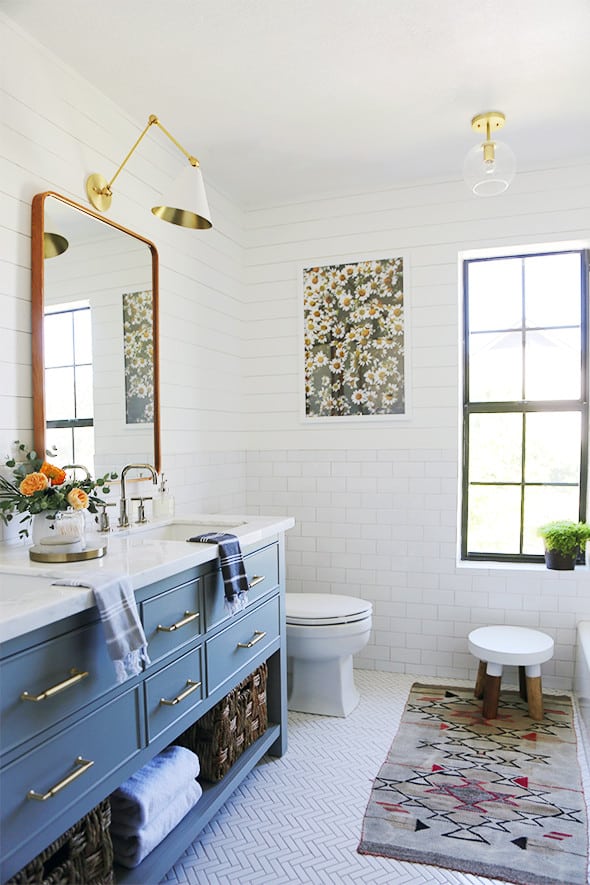 This light and bright modern vintage bathroom combines subway tile and shiplap on the walls. The double vanity provides a pop of color with navy blue cabinets and gold hardware. A wood-framed mirror hangs above the sinks.