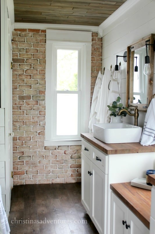 I love the vintage exposed brick wall in this otherwise modern style bathroom. The white vanity has butcher block style countertops and cabinets with dark polished brass hardware. The natural tones of the brick are echoed in the wooden countertops, wooden mirror frame, and vintage brass light fixtures.