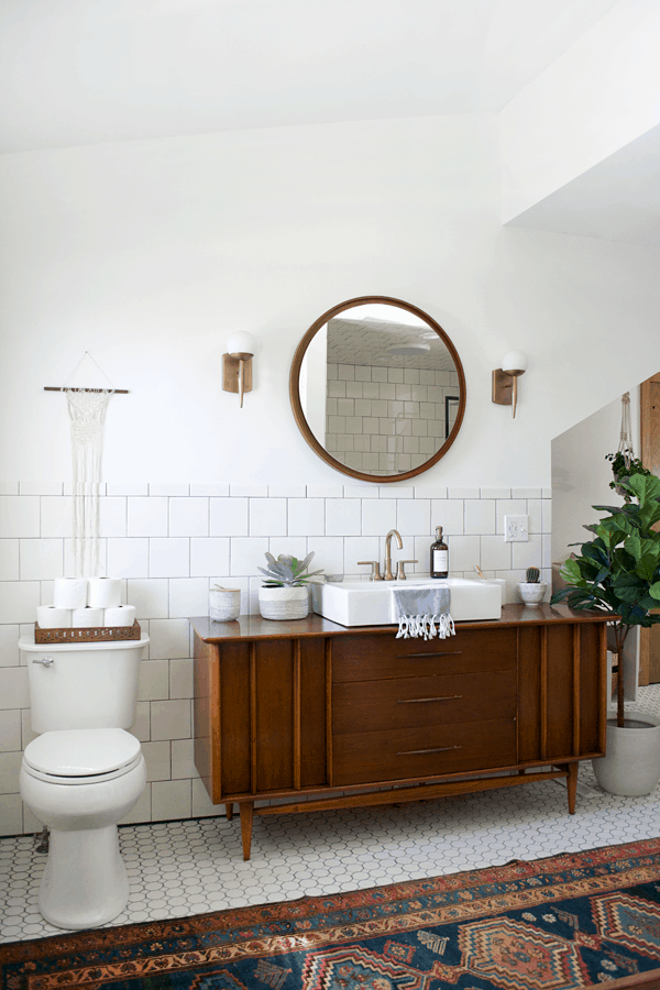 This modern vintage style bathroom is primarily white with a dark wood  vintage dresser converted into a vanity with sink. Round tiles line the floor, and white square tiles line the walls. A wood-framed round mirror hangs above the vanity.