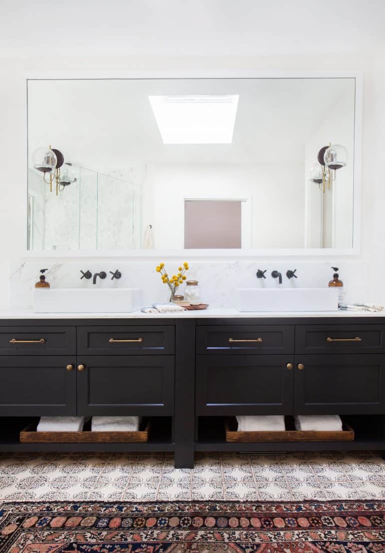 This bright and airy bathroom balances light and dark with a large double sink vanity with black cabinets and white marble countertops and backsplash. The large white-framed mirror is accented by vintage globe light sconces.