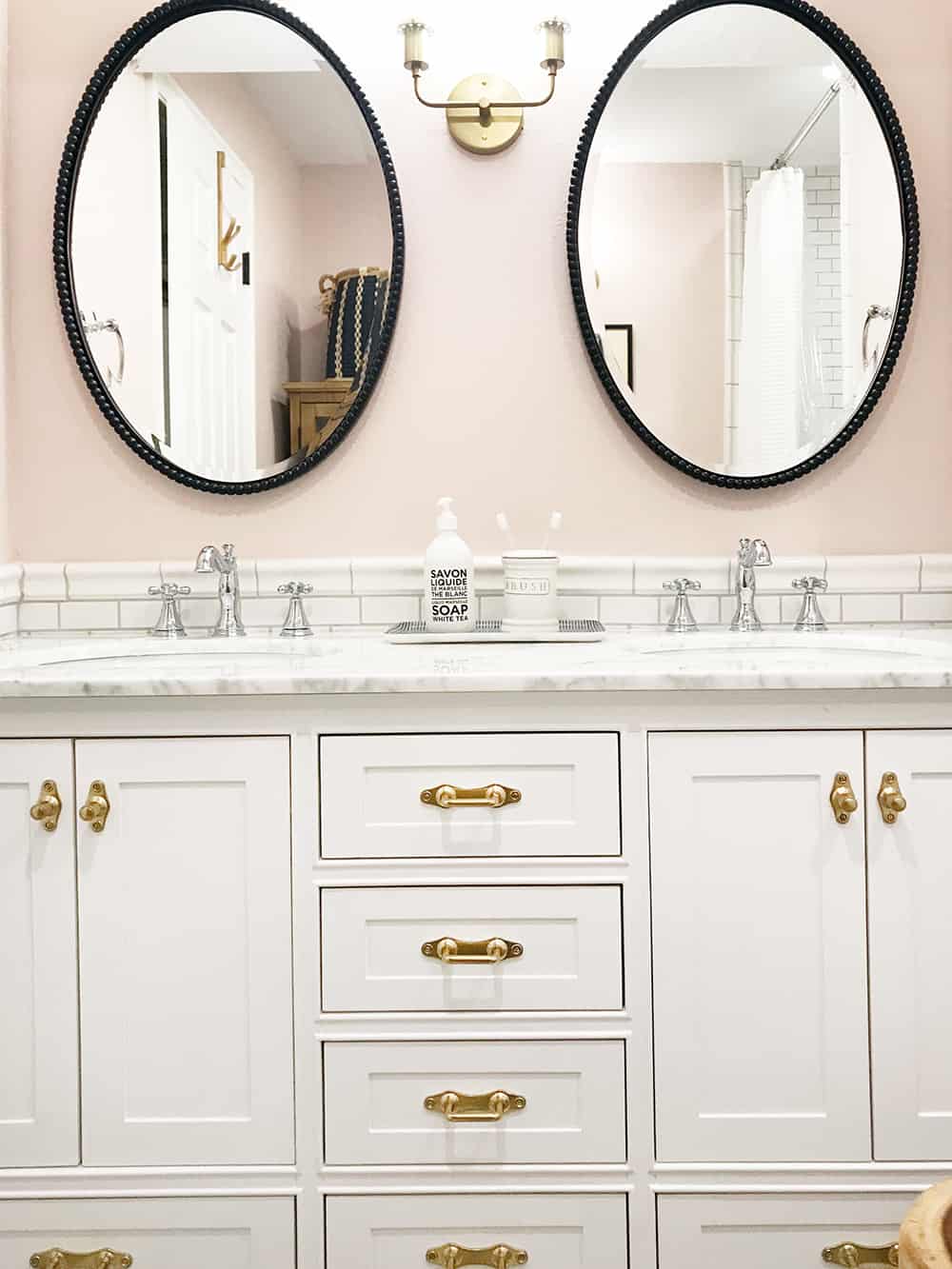 A double-sink bathroom vanity with white cabinets and white marble countertops. the bathroom fixtures are mixed metals - gold cabinet handles, chrome faucets, and black metal framed oval mirrors handing above each sink.