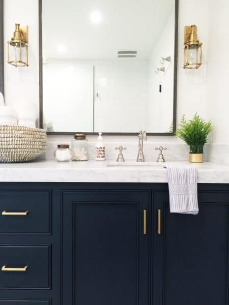 A bathroom vanity dark black painted cabinets and white marble countertops. Mixed metals throughout the vanity are brass cabinet handles, polished chrome sink faucet, brass sconces, and a metal-framed hanging mirror.