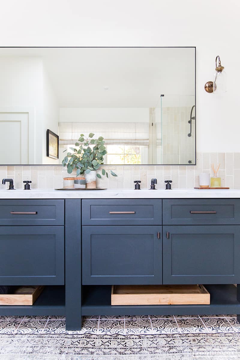 A double bathroom vanity with navy blue cabinets and mixed metal fixtures. A large rectangular mirror hangs on a white wall, reflecting back a window and glass shower.