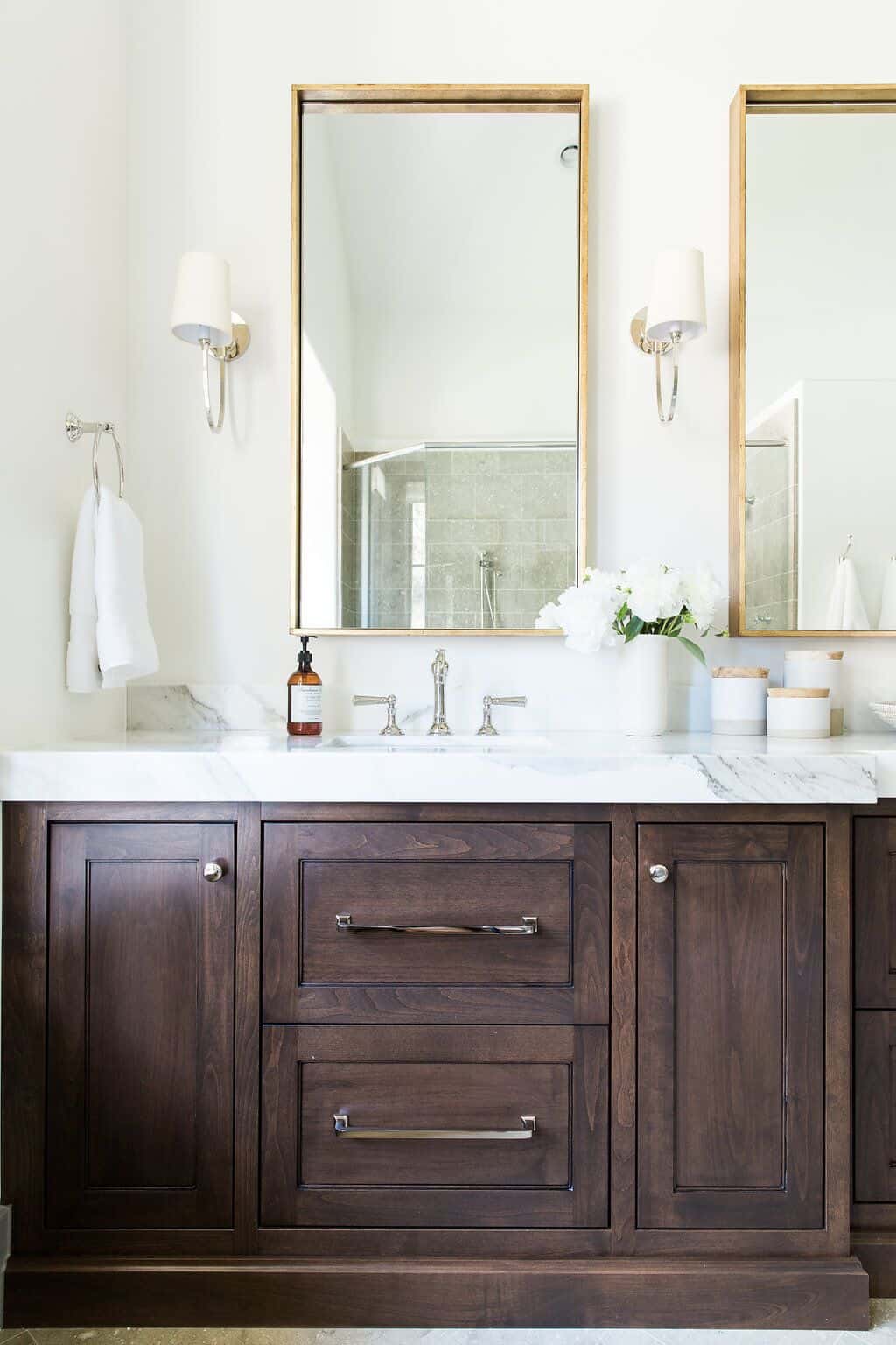 A bathroom vanity with dark brown wood cabinets and marble countertops. The fixtures in the bathroom are mixed metals - polished chrome faucets, chrome cabinet handles, and fold-framed mirrors.