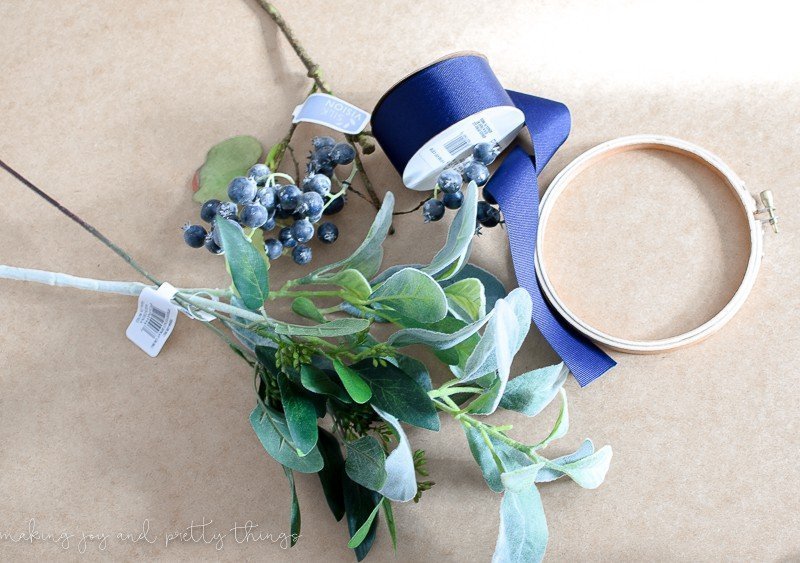 All the supplies needed to make a DIY embroidery hoop wreath, laid out on a brown surface. Steps of lamb's ear leaves, faux berries, navy blue ribbon, and a plain wood embroidery hoop.