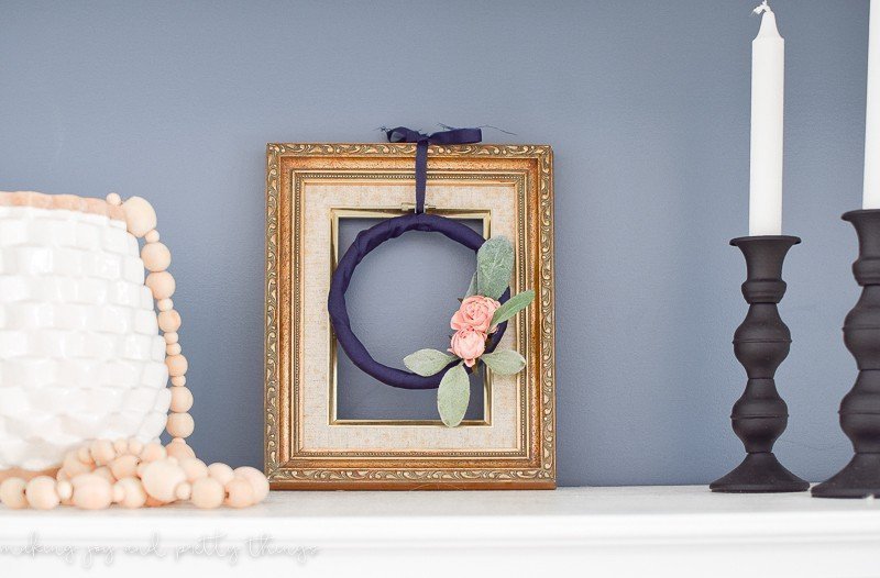 A small embroidery hoop wreath made with navy blue ribbon, blush-pink roses, and lamb's ear hangs in a white and gold picture frame on a marble surface. To the left is a white vase with wood bead garland; to the left are two black candlesticks with white pillar candles.