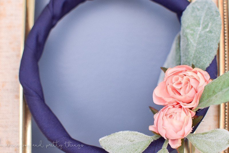 A close up look at the details of the DIY embroidery hoop wreath - navy blue fabric is wrapped around a wood embroidery hoop with two mini blush-pink faux roses and faux lambs ear leaves.