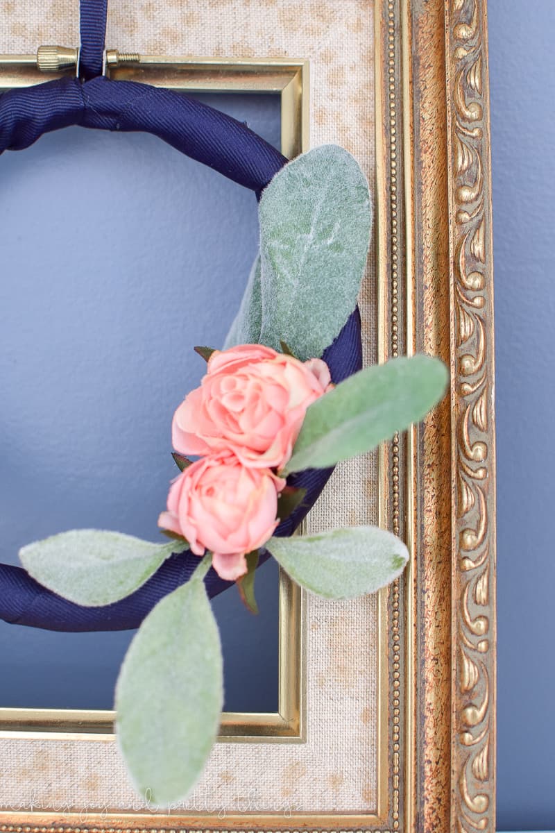A close up look at the details of the embroidery hoop wreath and thrifted picture frame. The embroidery hoop wreath has navy blue fabric, faux lamb's ear leaves, and blush-pink roses. The frame has an ornately detailed gold-painted wood frame.