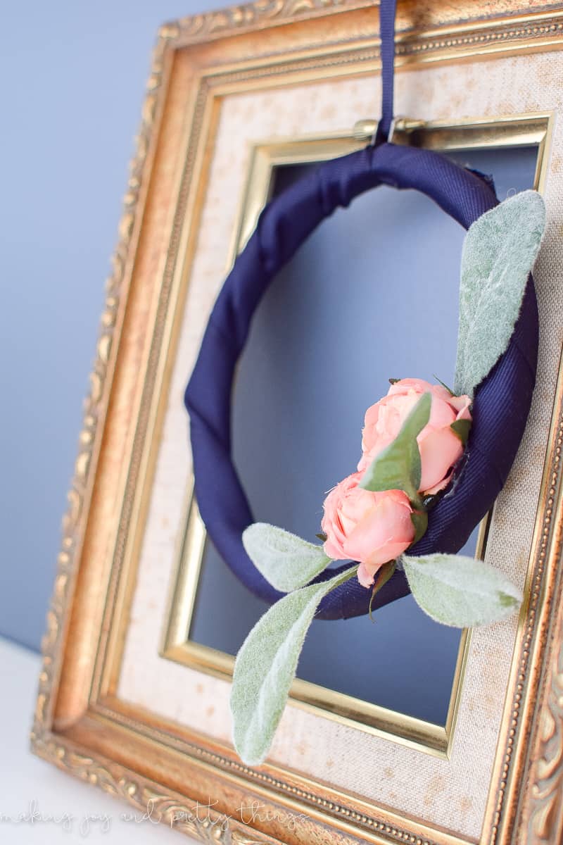 A side view of the embroidery hoop wreath hanging in a thrifted gold-painted picture frame. The wreath is made from a navy blue fabric-wrapped embroidery hoop with blush pink roses and lambs ear leaves.