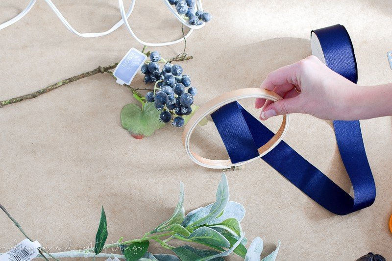 A woman's hand holds a wooden embroidery hoop frame; the end of the navy blue ribbon is attached to the inside of the hoop with glue. The other embroidery hoop wreath supplies - steps of faux flowers and leaves - sit alongside the hoop.