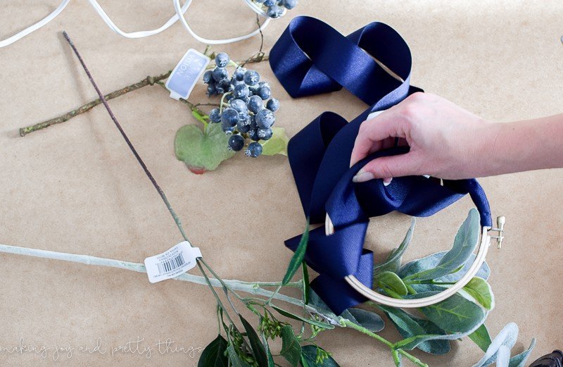 A woman's hand wraps a wide navy blue ribbon around a wooden embroidery hoop frame. Stems of faux flowers, berries, and leaves sit on the table around the craft.
