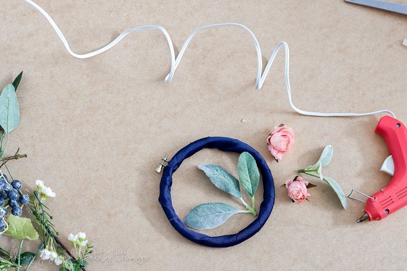 An embroidery hoop sits wrapped with navy blue fabric. Two mini pink rose heads are detached from the stems, sitting next to a glue gun, which is plugged in and warming up. On the left side of the image, more stems of faux flowers sit, partially visible.