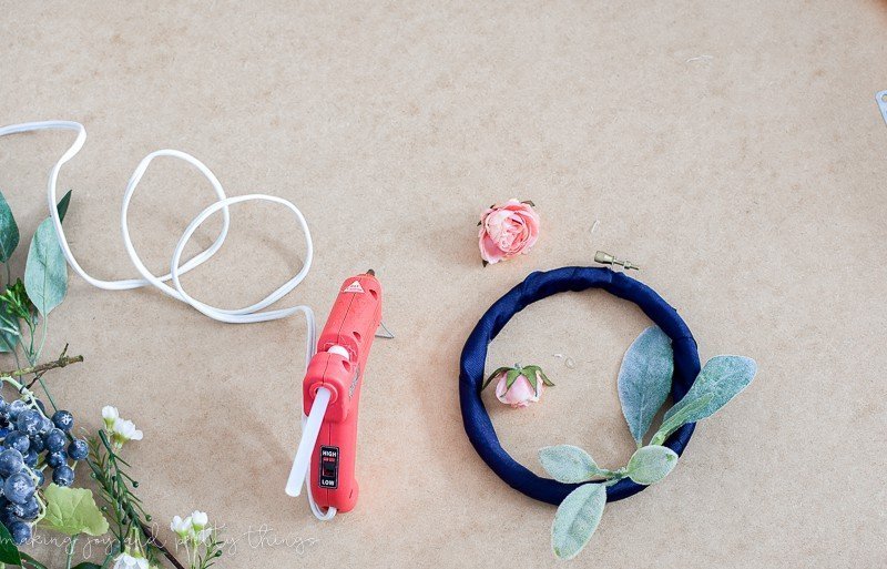 Five faux lamb's ear leaves are glued to a navy blue ribbon-wrapped embroidery hoop, and an orange glue gun sits on the table next to the hoop, plugged in with a white cord. Two light pink rose heads sit detached from their stems, next to the embroidery hoop wreath.