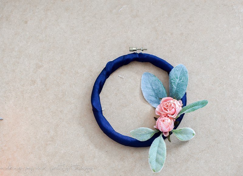 A completed embroidery hoop wreath wrapped with navy blue ribbon and decorated with faux lamb's ear leaves and two blush-pink mini roses.