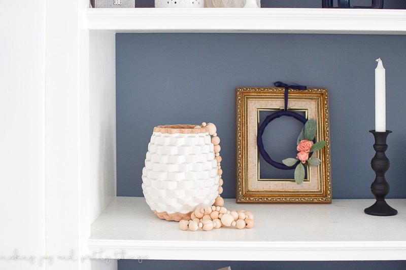 The embroidery hoop wreath and picture frame sits on a white bookshelf alongside a white vase filled with a wood bead garland and a black candle stick with white pillar candle.