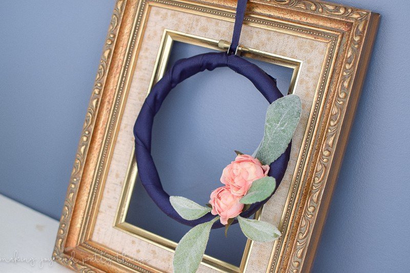 A closer look at the DIY embroidery hoop wreath, made with navy blue ribbon and decorated with mini blush-pink roses and faux lamb's ear. The wreath hangs in the center of an empty picture frame with a stylish gold finish.