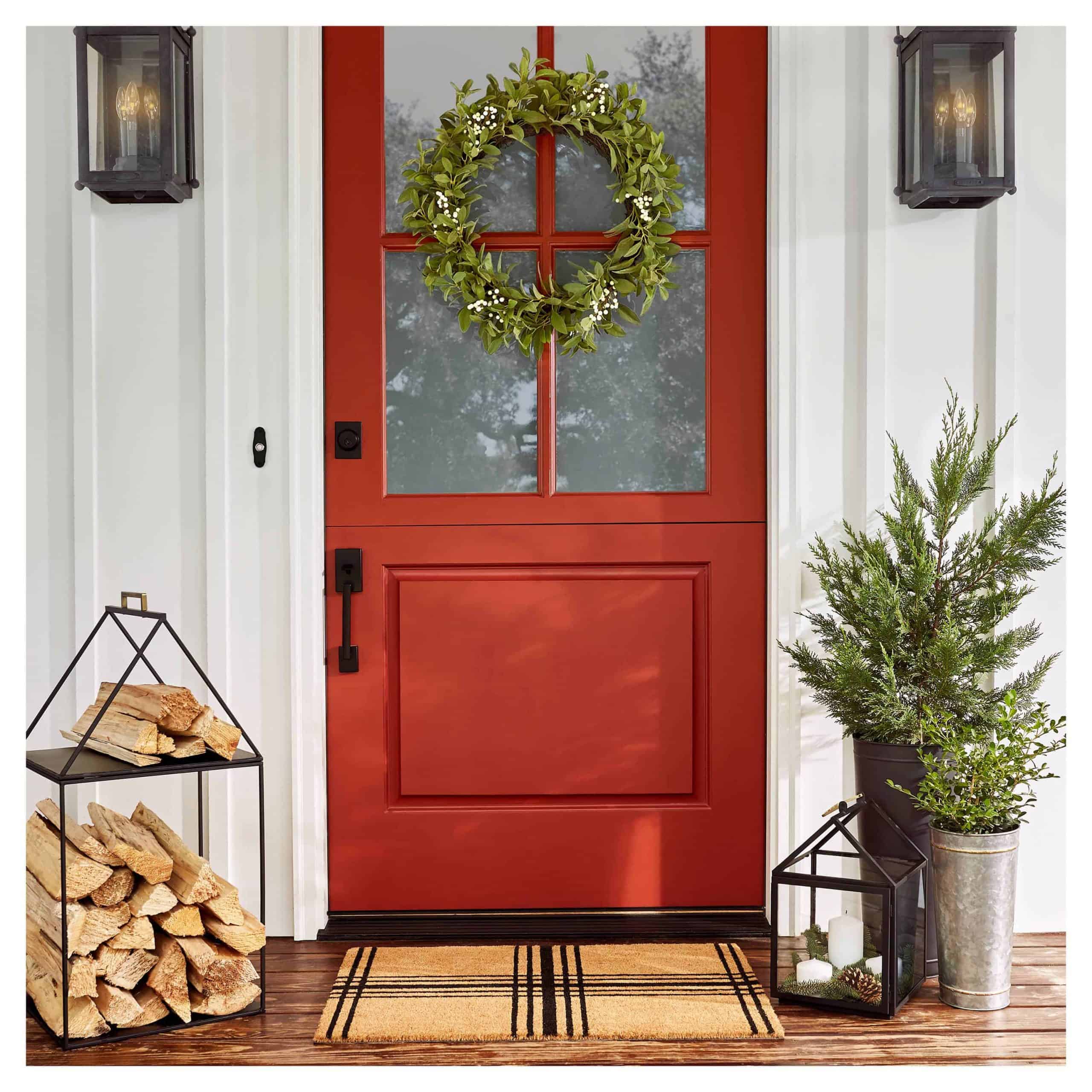 A photo of a red door with a wreath hanging on it and chopped wood and plants on the side.
