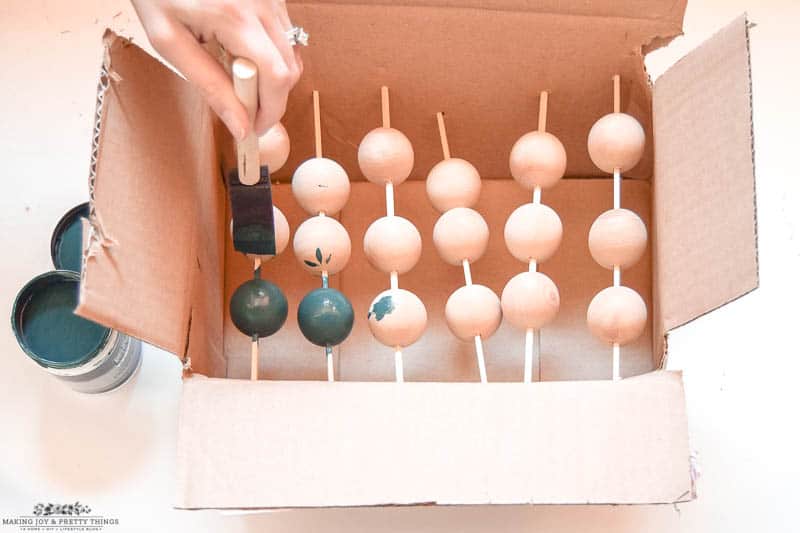 Painting oversized wood bead garland with a green paint to completely cover and coat wooden balls with sticks in-between