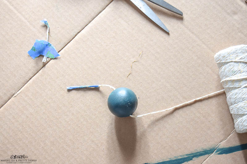 Showing how to thread a bead with white and gold twine that has been taped on end on a carboard box