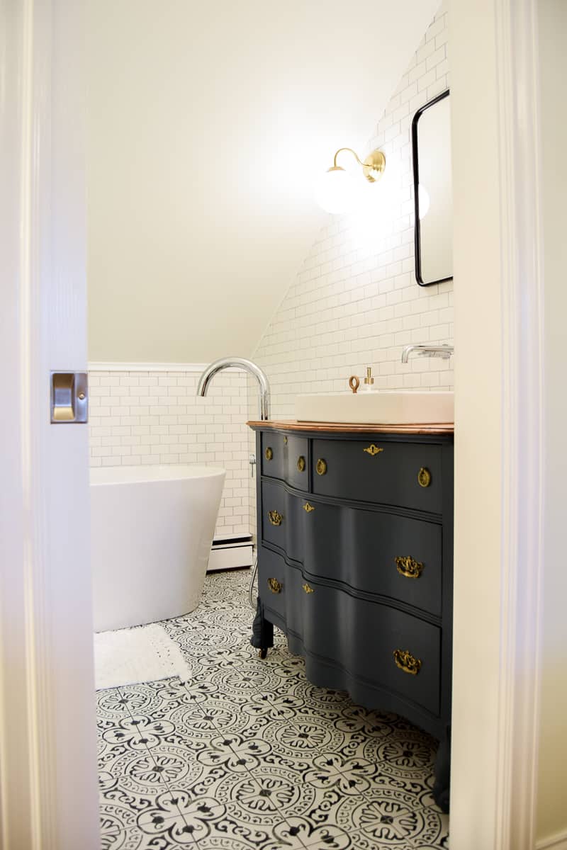 The completed modern vintage bathroom reveal! The view from the bathroom door shows the vintage sink vanity, painted black with gold hardware, the deep-set bathtub, and white subway tile walls.