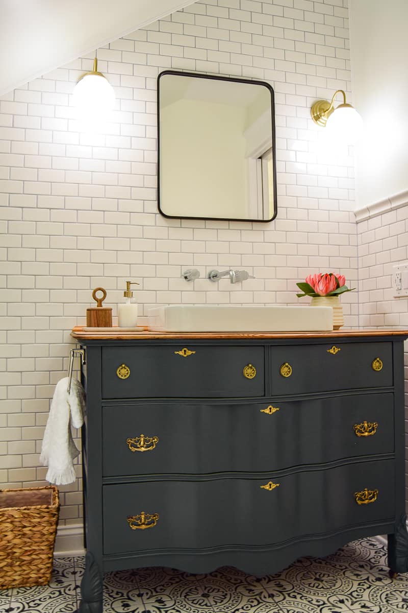A complete look at the modern vintage vanity set up in our bathroom: a vintage dresser repurposed into a vanity with fresh paint, inlayed porcelain sink, and silver wall-mounted fixtures. Brass globe wall scones hang on either side of a sleek black-framed bathroom mirror.