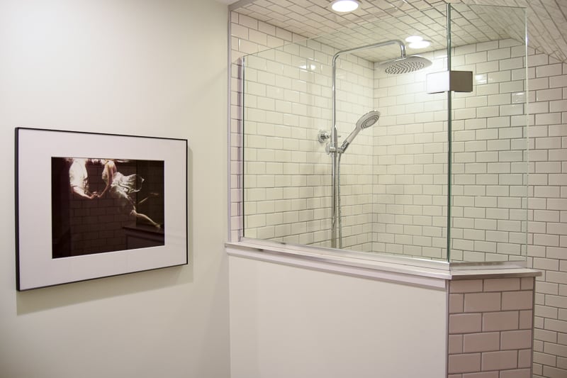 A gorgeous walk-in shower with white subway tiled walls and glass window, rainfall showerhead, and framed photography on the white painted walls.