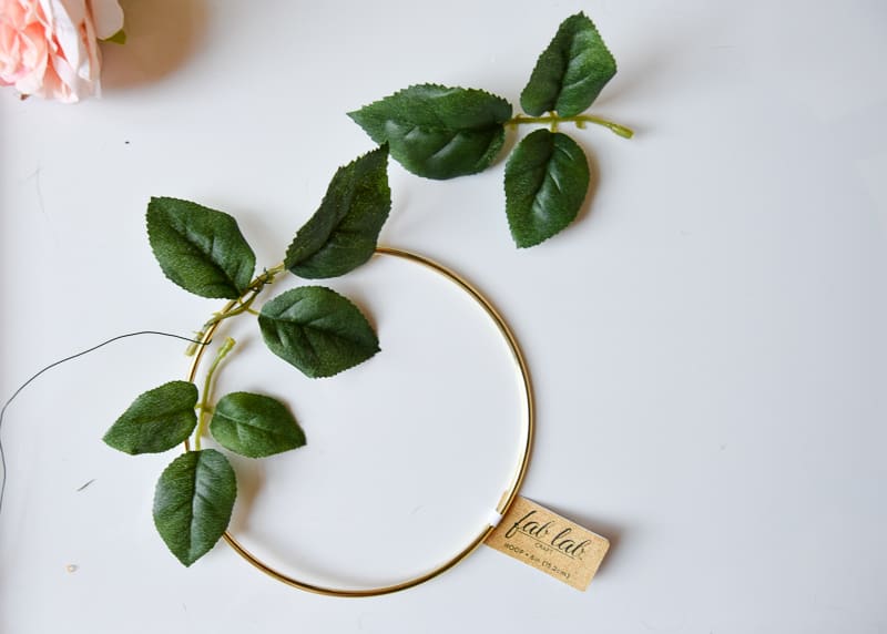 Sprigs of faux green leaves are wrapped around a gold macramé hoop with floral wire.