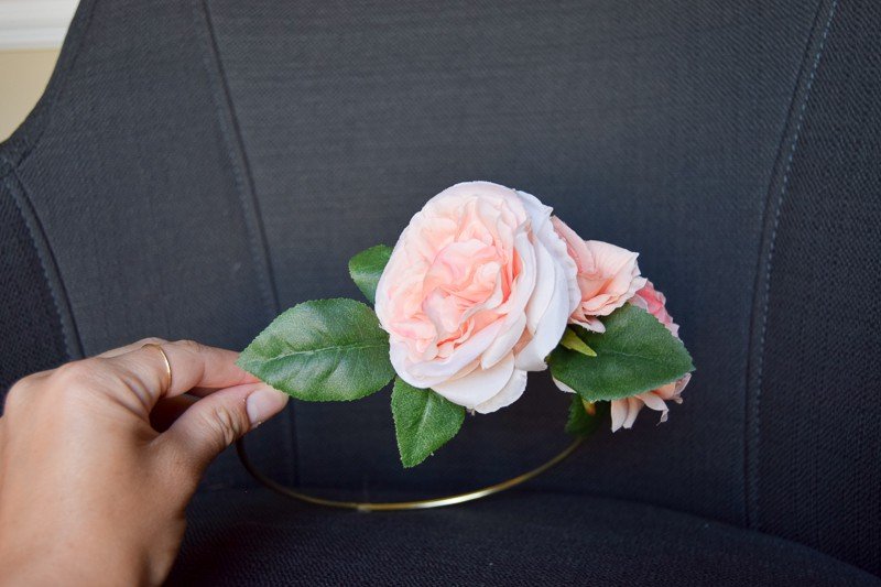 A woman's hand holds the finished floral crown in front of a denim fabric chair. The floral crown has three pink peony flowers and green leaves attached to a gold hoop.