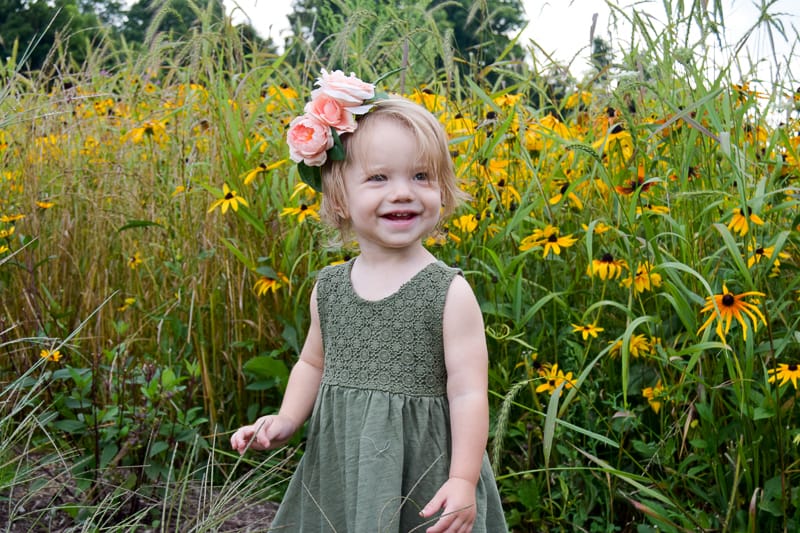 A little girl stands in front of a field of tall grass and yellow wild flowers. She's wearing an olive green dress and a flower crown made with faux pink peony flowers and leaves.