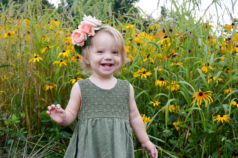 A little girl in an olive green dress smiles at the camera. She's wearing a pink peony floral crown and is standing in front of a field of tall grass and yellow wild flowers.