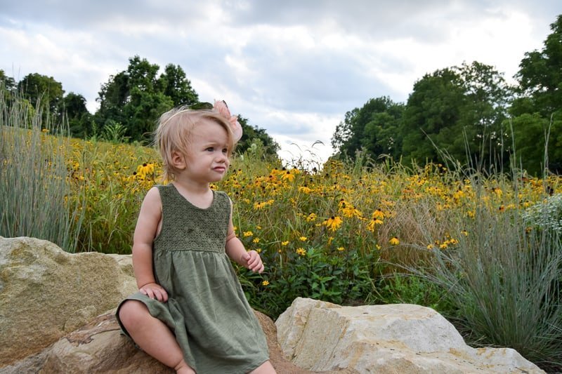 A little girl sits on some rocks in front of a field of grass and yellow wild flowers. She's wearing a green dress and a DIY flower crown made from pink peony flowers. She had a slightly worried expression on her face, like she's about to cry.