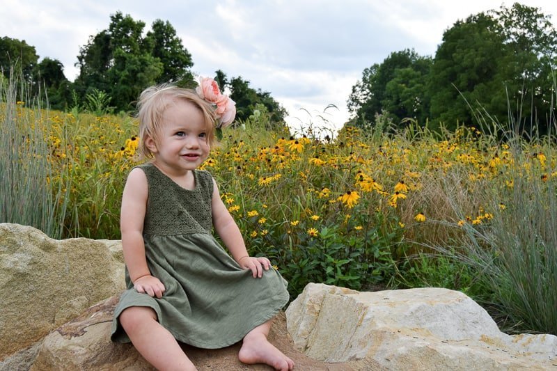 A little girl in a green dress sits on some rocks in front of a field of tall grass and yellow wild flowers. She's wearing a pink peony flower crown on her head and smiling at the camera.