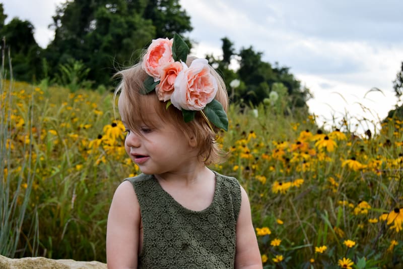 A little girl in an olive green dress looks over her shoulder. She's wearing a DIY flower crown made with pink peony flowers and leaves, and is standing in front of a wild flower field with yellow flowers.