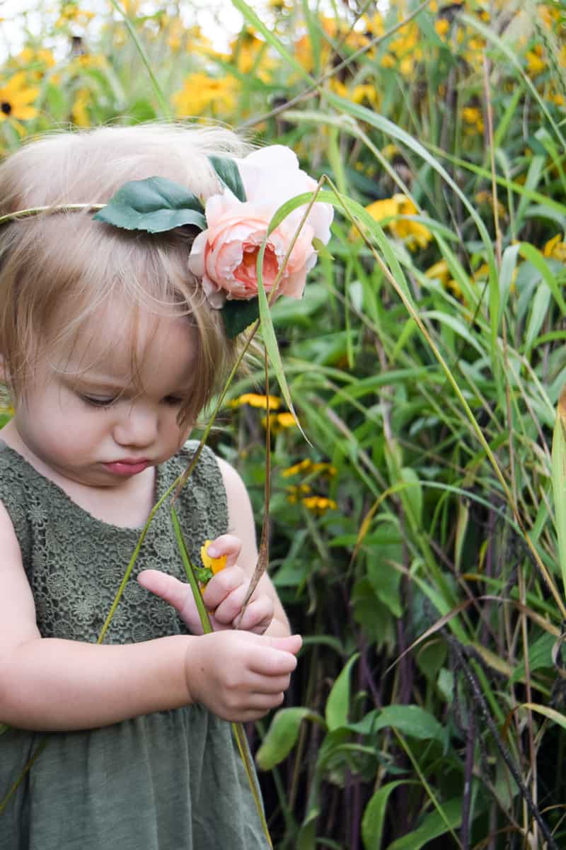 A little girl wearing an olive green dress plays with tall blades of grass and flower petals in a grassy field. She's wearing a flower crown made with pink peony flowers and leaves on her head.