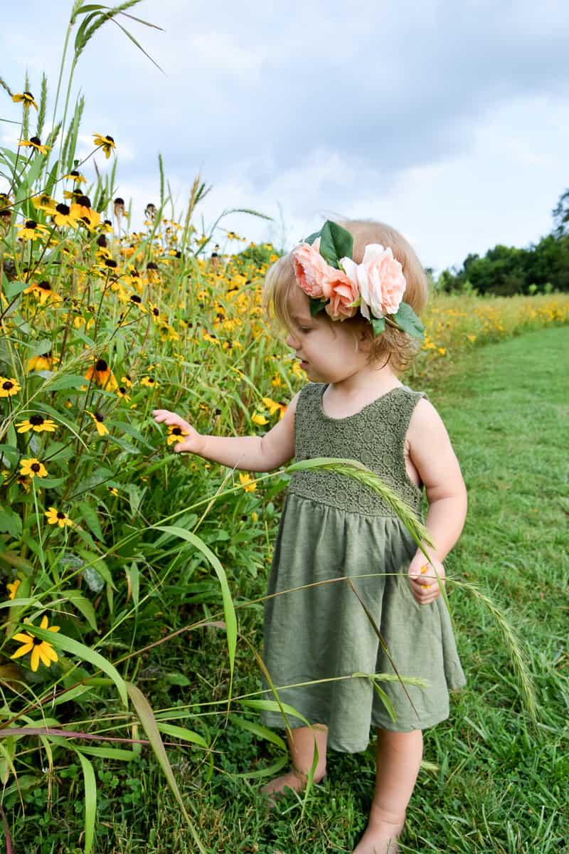 A little girl wearing an olive green dress stands in front of a field of tall grass and yellow wild flowers. She's playing with the flowers in front of her. On her head, she's wearing a DIY flower crown made with pink peony flowers and leaves.