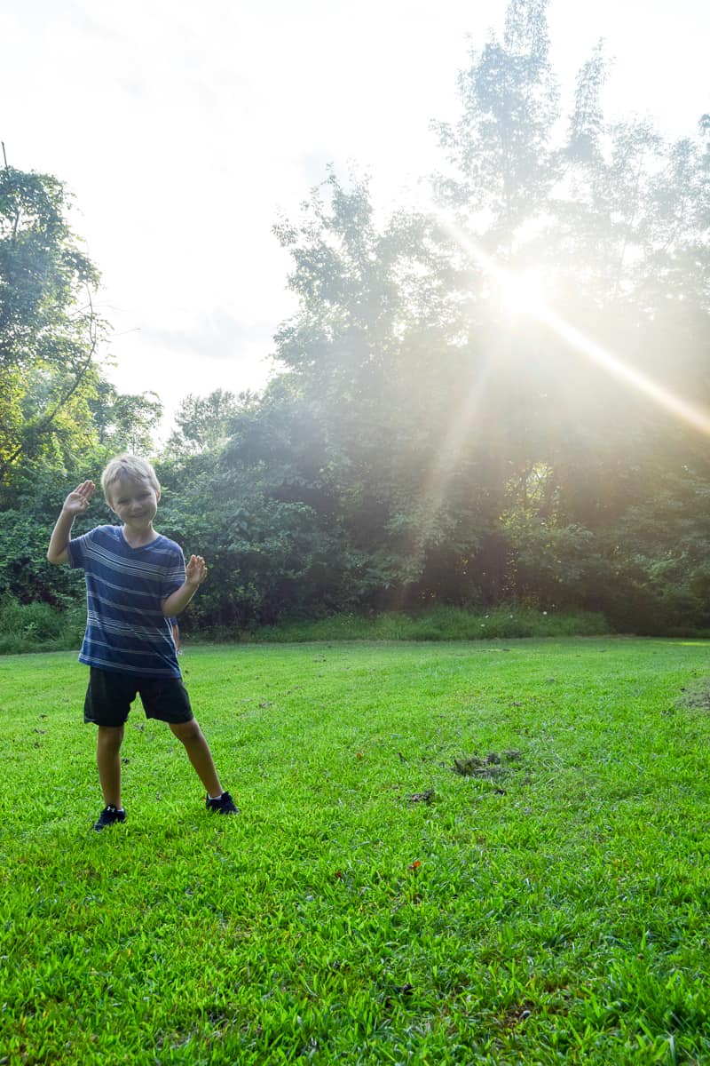 A young boy stands in a grassy field with woods behind him. He's wearing shirts and a stripped blue top and smiling and waving at the camera. 