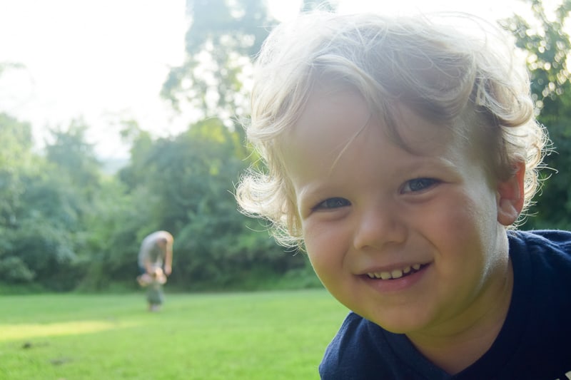A little boy's face is close to the camera, smiling a sly grin. In the background, a little girl and an adult are out of focus in the field.