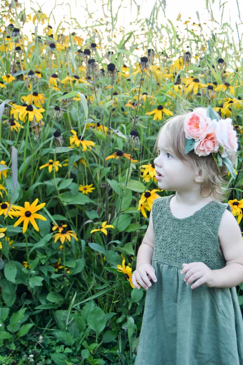 A little girl stands in front of a field of tall grass and yellow wild flowers. She's wearing a green dress and a pink peony flower crown, looking out onto the flower field.