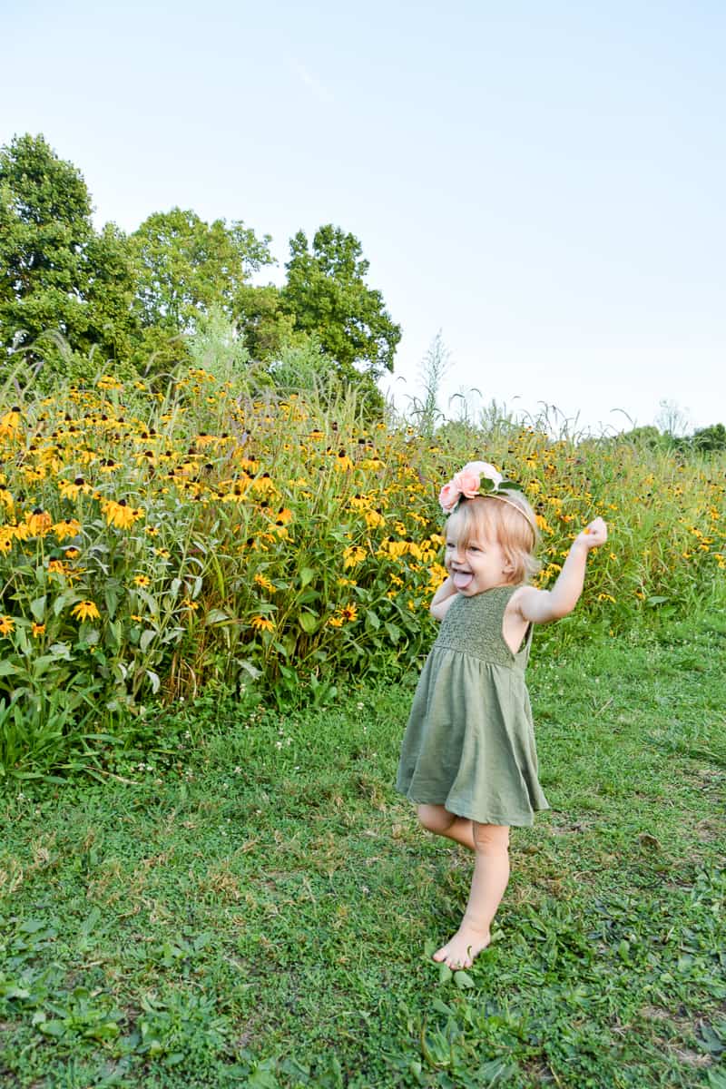 A little girl in an olive green dress smiles with her tongue out while dancing in a grassy field with wild flowers. She's wearing a DIY flower crown on her head, made with pink peony flowers and leaves.