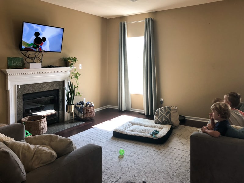 A plain living room with dark tan walls and minimal natural light. Kids sit on a couch, watching TV, which is mounted on the wall over a white and marble fireplace. A large white area rug covers most of the dark hardwood floors.