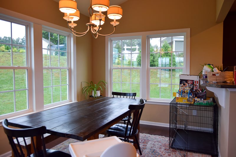 Our old dining room before the makeover. Large windows line the beige walls. A large natural wood dining room table with chairs sits under and outdated chandelier. A large dog crate is tucked in the corner of the room.