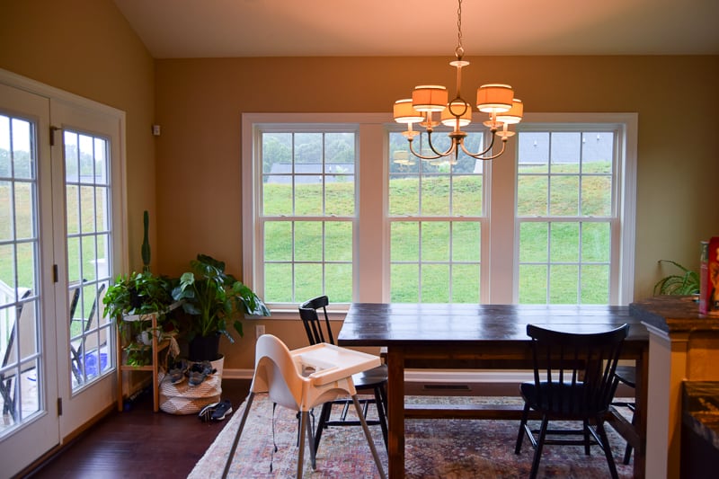 A dining room with large windows and doors, beige walls, and natural light. An outdated chandelier hangs over a large natural wood dining table, surrounded by chairs and a baby's high chair.